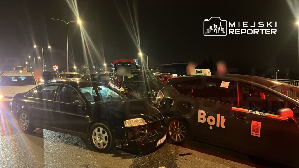 Zderzenie dwóch samochodów osobowych na ruchliwej ulicy nocą, w tle widoczne inne pojazdy i latarnie.