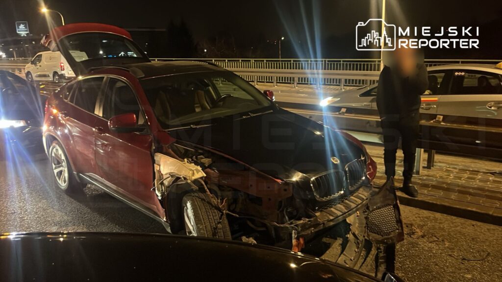 Czerwony SUV z uszkodzoną przednią częścią stoi na drodze w nocy, obok znajduje się mężczyzna w czarnym ubraniu.