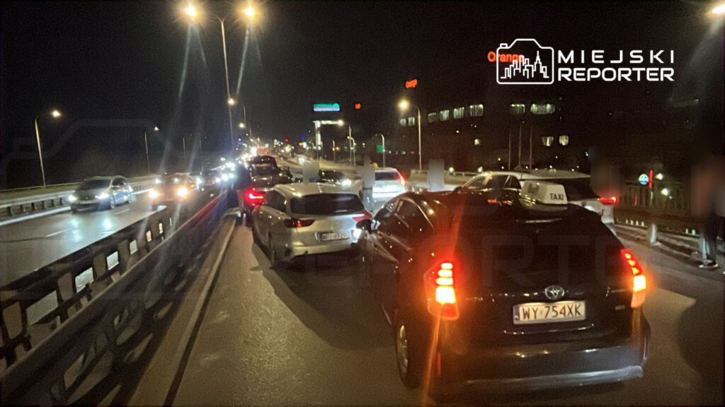 Ruch uliczny w nocy z samochodami stojącymi w korku na jezdni, w tle widoczne oświetlone budynki.