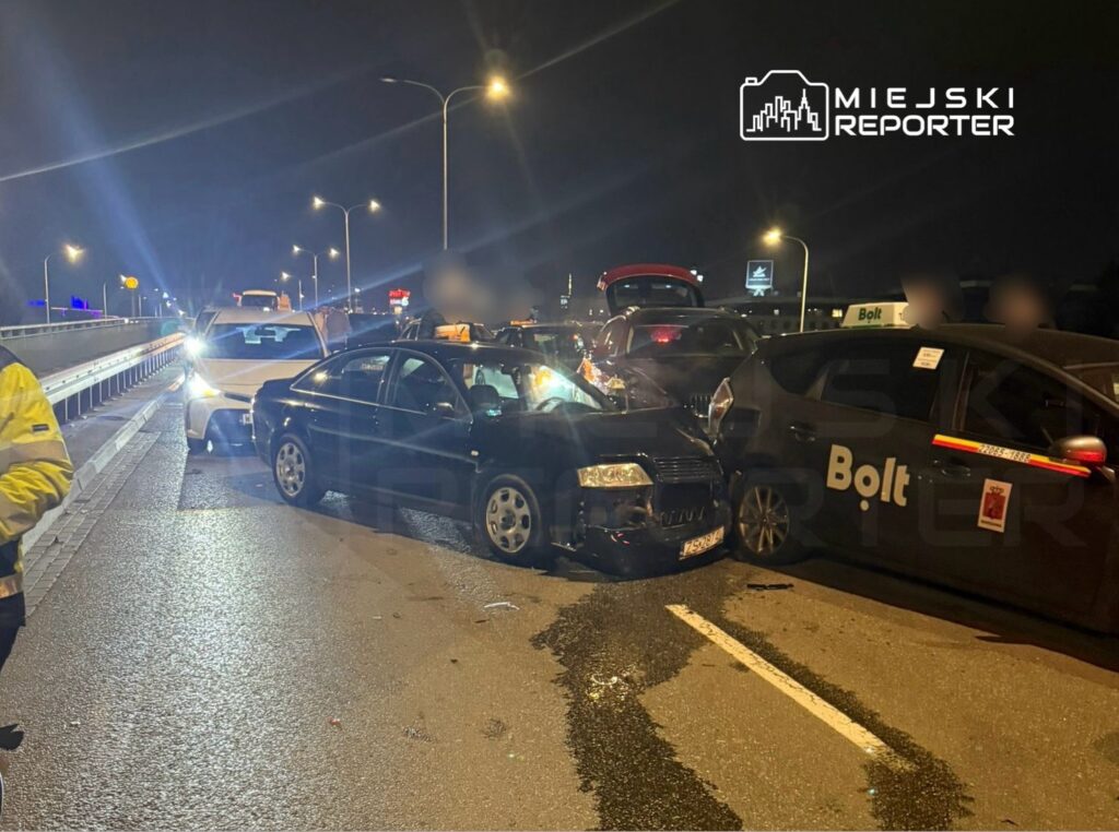 Zderzenie trzech samochodów na oświetlonej drodze nocą, z osobami na poboczu.