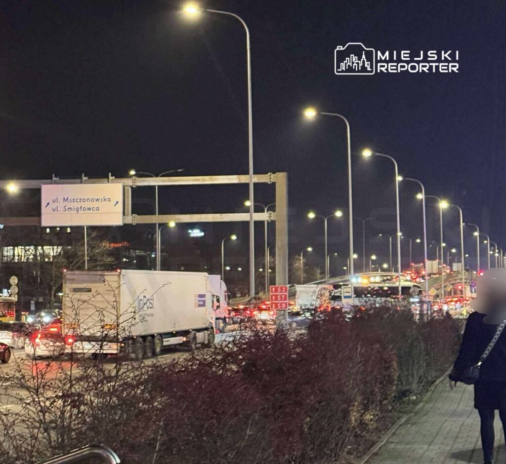 Ruchliwa ulica nocą, z dużymi truckami i samochodami osobowymi w korku, oświetlona latarniami. Widoczny znak drogowy.