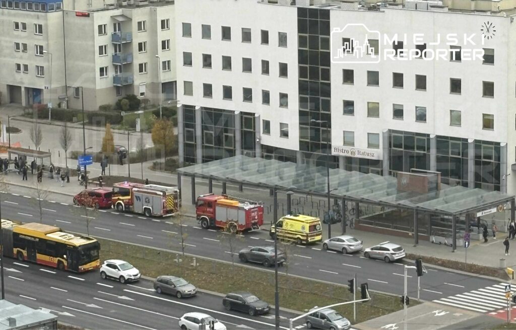 Na ulicy stoją dwa samochody strażackie oraz jeden ambulans, obok przystanku komunikacji miejskiej. W tle widać nowoczesny budynek z przeszkloną fasadą oraz napis "Bistro Katwusz". Przechodnie czekają na autobus, a w oddali widać ruch uliczny.