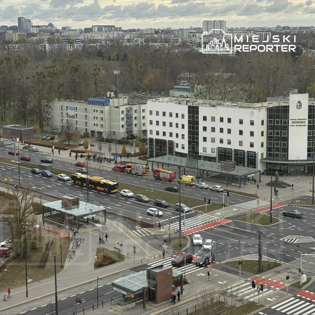 Na skrzyżowaniu ulic parkują pojazdy straży pożarnej, obok przystanku autobusowego w nowoczesnej zabudowie.