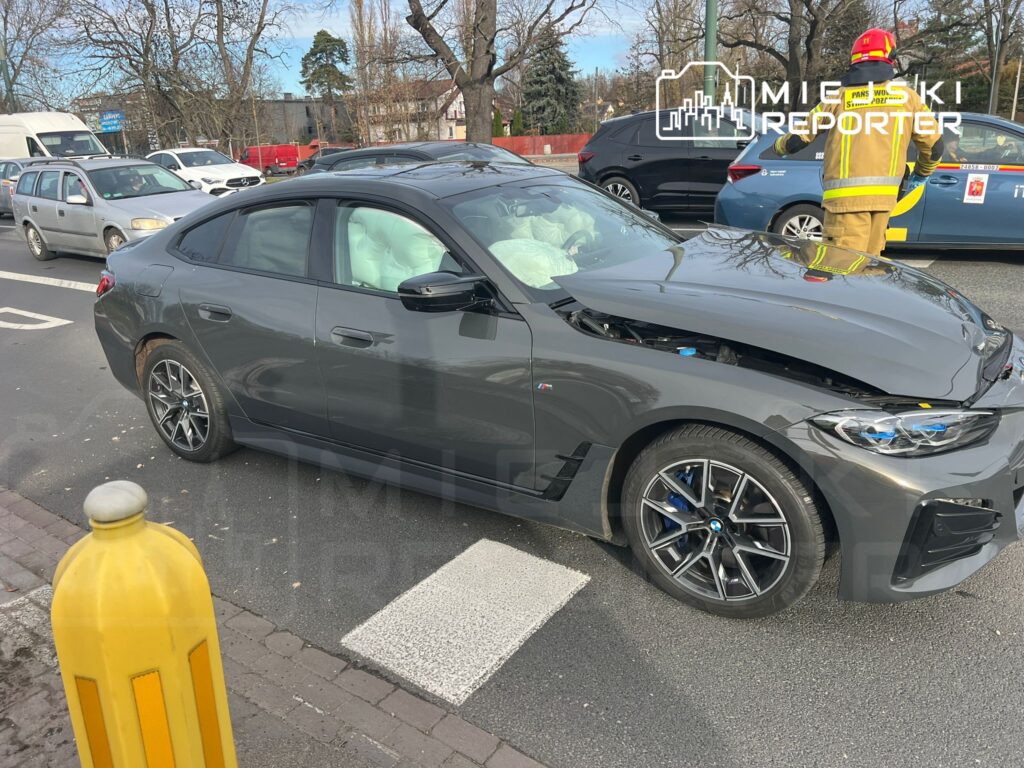 Czarny samochód osobowy z uszkodzoną maską stoi na drodze, obok interweniujący strażak w żółtym mundurze.