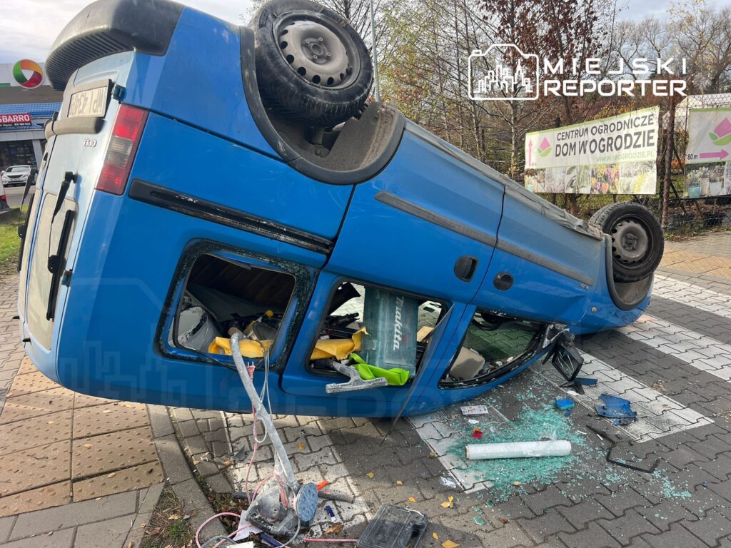 Niebieski furgon leży na dachu na chodniku, otoczony rozbitym szkłem i uszkodzonymi przedmiotami.