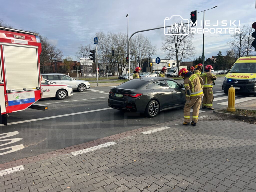 Grupa strażaków w mundurach i hełmach udziela pomocy przy uszkodzonym czarnym samochodzie na skrzyżowaniu.
