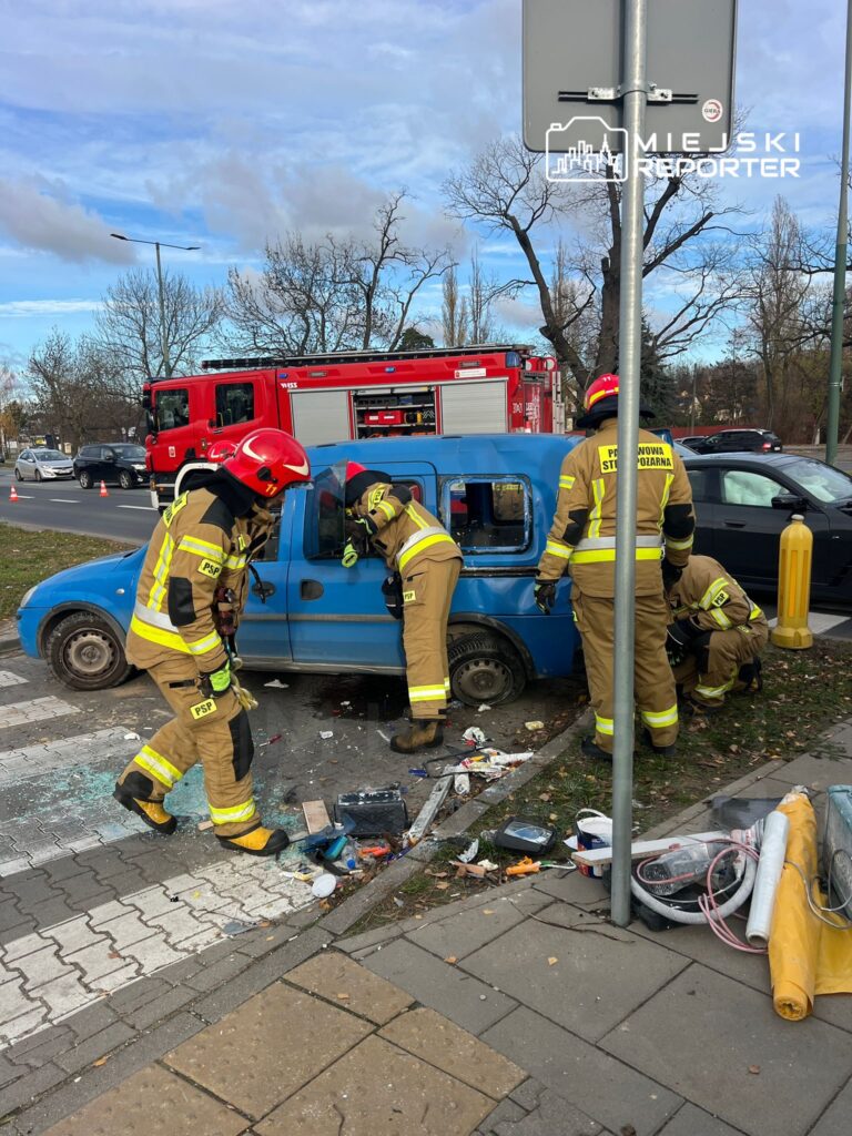 Czterech strażaków w mundurach pracuje nad niebieskim samochodem osobowym, który wjechał w przeszkodę. W tle widać wóz strażacki.