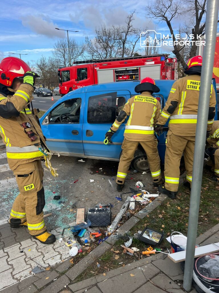 Strażacy w mundurach ratunkowych pracują nad wydobyciem osoby z niebieskiego pojazdu osobowego na chodniku.