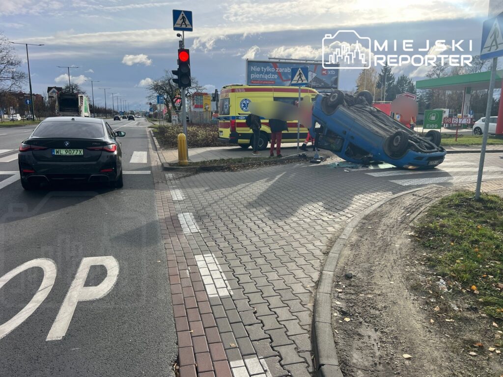 Na skrzyżowaniu leży przewrócony niebieski samochód, obok niego stoją ratownicy medyczni i karetka. W tle widoczny czarny samochód osobowy.