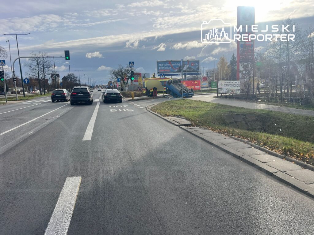 Na skrzyżowaniu z zielonym światłem stoją trzy samochody, a obok leży przewrócony pojazd. W tle widoczne reklamy i znak "DOP BUS".