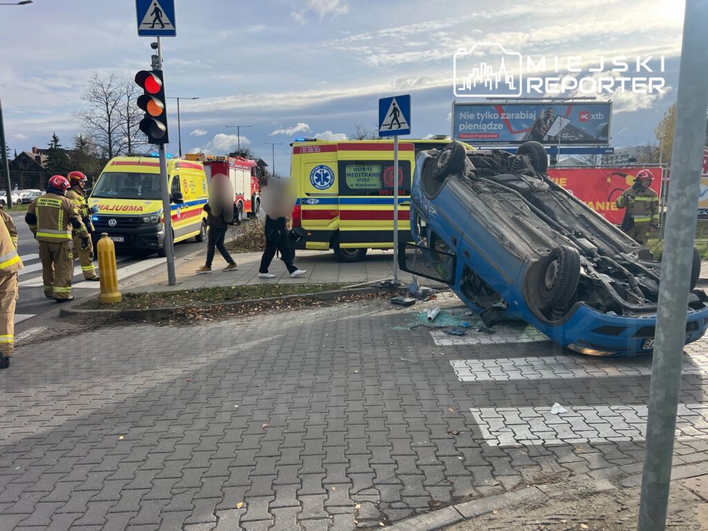 Nieoznakowany niebieski samochód leży na dachu na skrzyżowaniu, wokół grupka strażaków i karetka pogotowia.