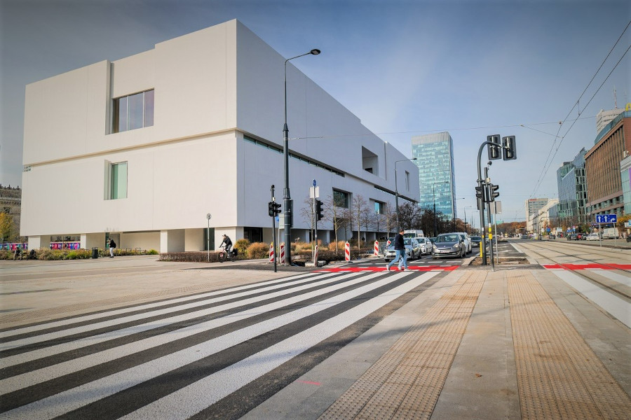 Nowoczesny budynek z białą fasadą znajduje się przy przejściu dla pieszych, obok ruchliwej ulicy i tramwajów.