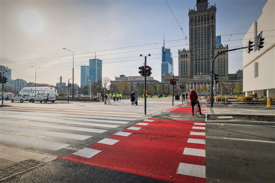 Ruchliwa ulica z przejściem dla pieszych, na którym stoi rowerzysta, w tle widoczny wysoki budynek.