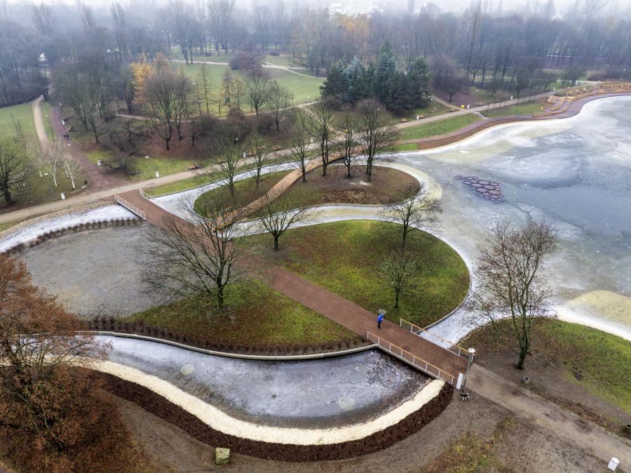 Widok na park z krętymi alejkami, mostkiem i zamarzniętym stawem otoczonym drzewami.