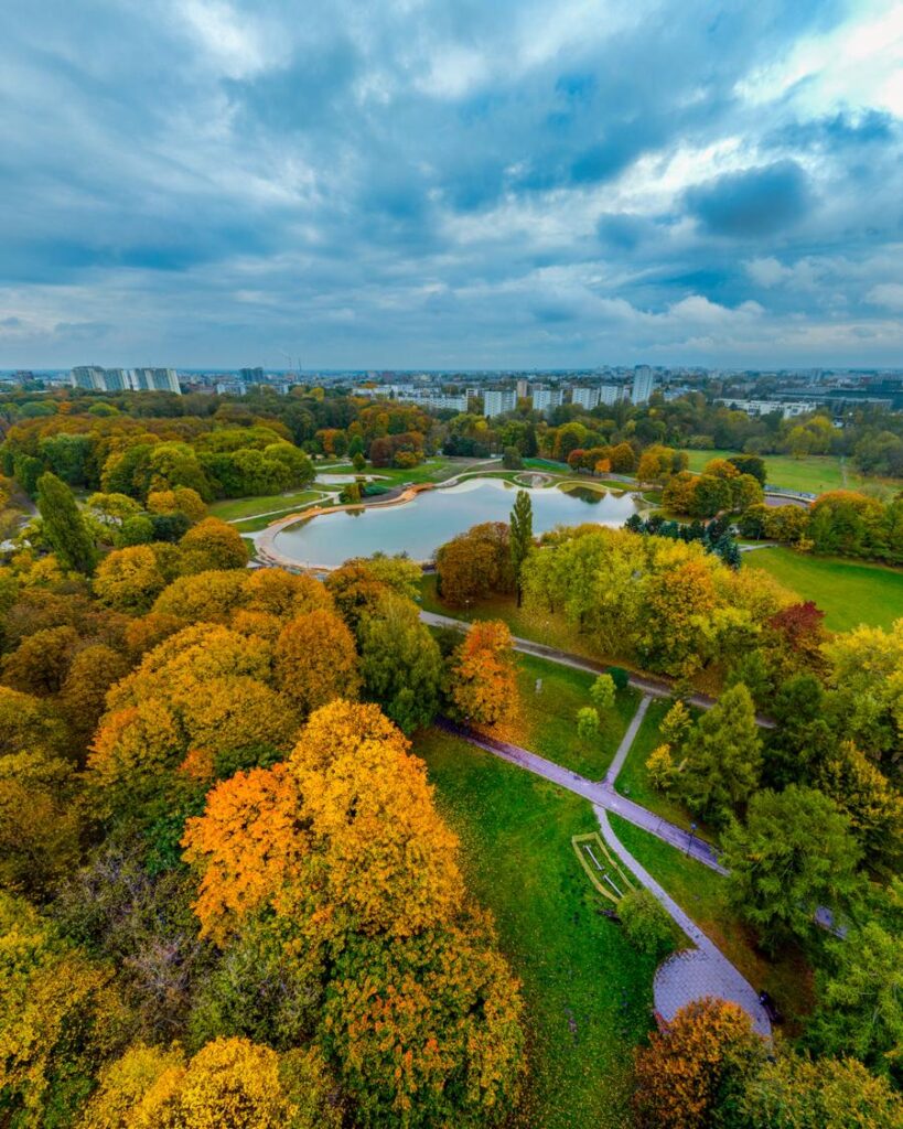 Widok na park z drzewami w jesiennych barwach oraz staw, w tle nowoczesne budynki i chmurne niebo.