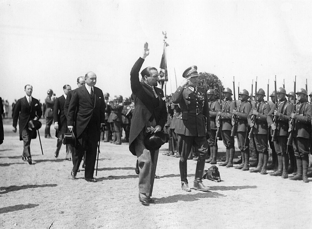 Mężczyzna w garniturze macha ręką, przechodząc obok żołnierzy w mundurach, którzy stoją na baczność.