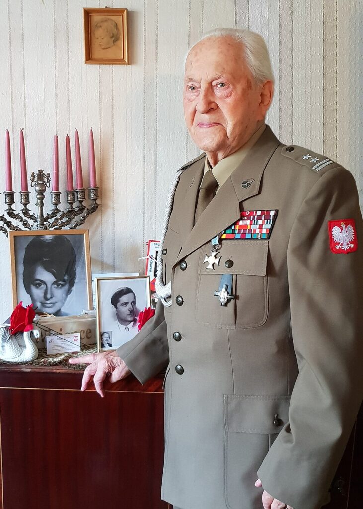 Starszy mężczyzna w mundurze wojskowym stoi obok stołu z pamiątkami, na którym znajdują się zdjęcia i świeczniki.