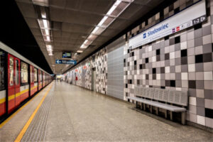 Pusta stacja metra z nowoczesnymi, szaro-białymi kafelkami i oznaczeniem "Słodowiec" na ścianie.