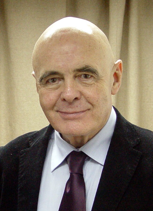 Mężczyzna z łysą głową, uśmiechający się w eleganckim ubraniu, na tle jasnej zasłony.