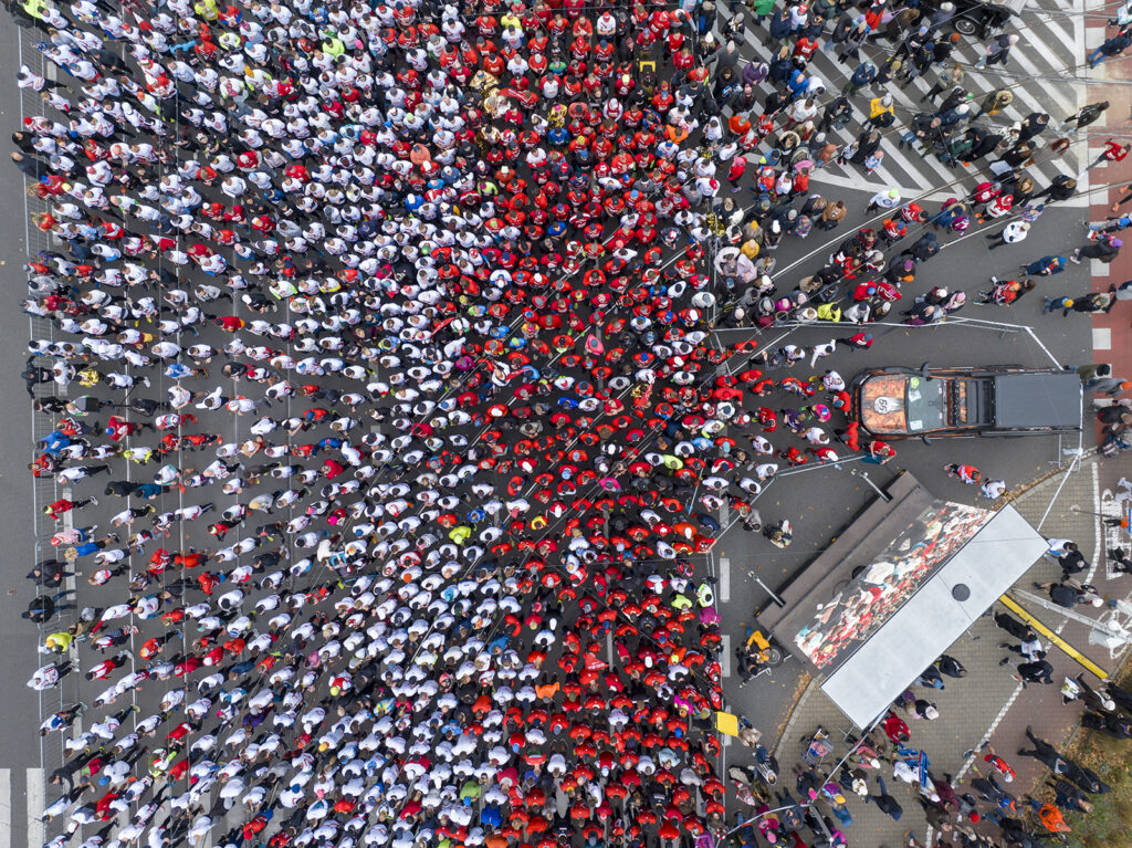 Tłum ludzi ubranych w czerwone i białe koszulki gromadzi się na ulicy, z samochodem i sceną w tle.