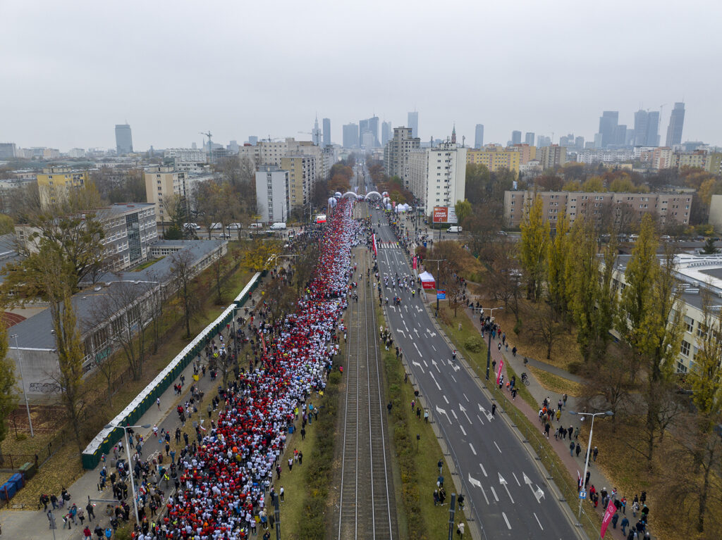Tłum ludzi w czerwonych i białych koszulkach maszeruje wzdłuż ulicy, otoczeni nowoczesnymi budynkami.