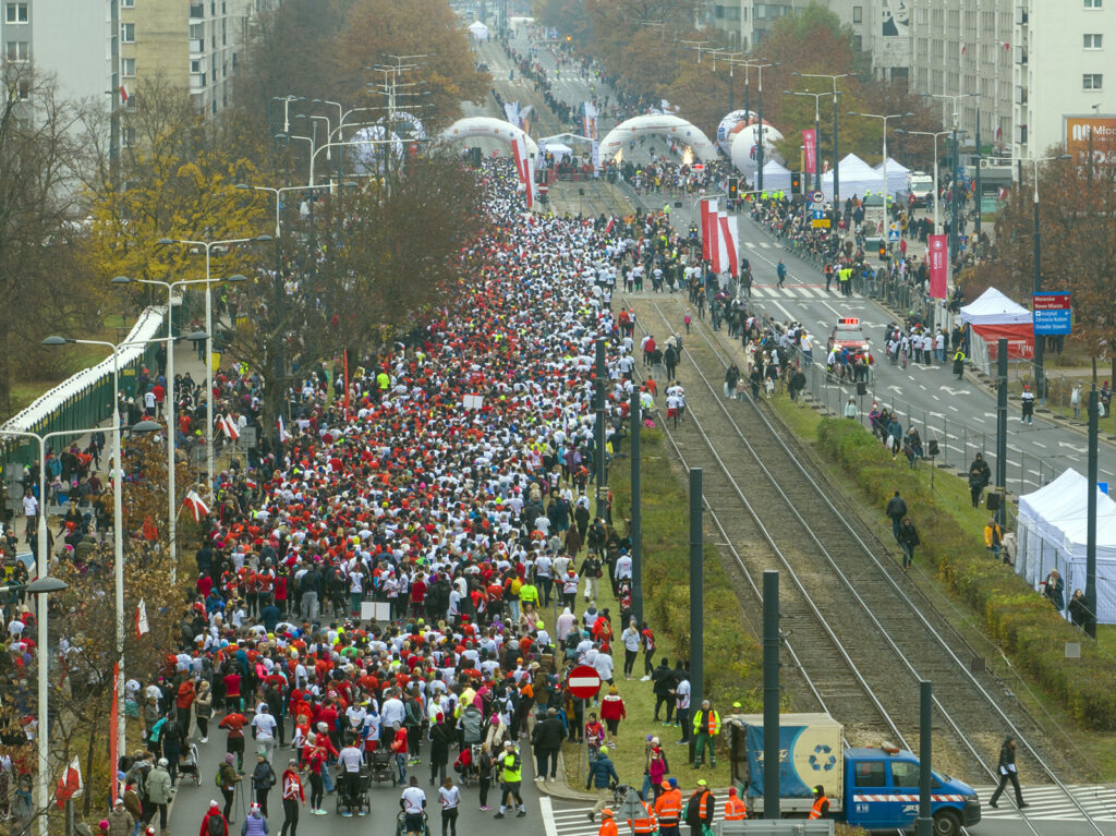 Tłum ludzi w białych i czerwonych koszulkach maszeruje ulicą, obok torów tramwajowych, w otoczeniu stoisk i flag.