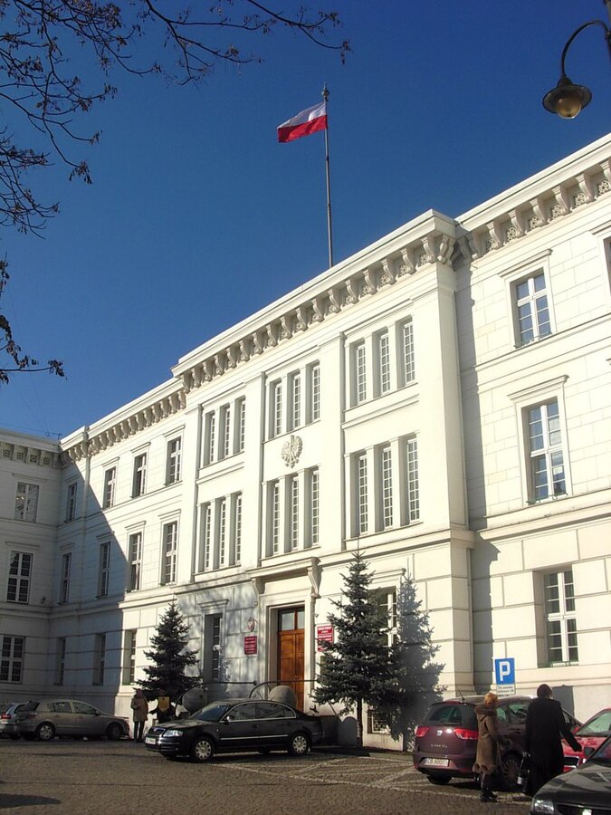 Biały budynek z flagą Polski na dachu, z samochodami zaparkowanymi na placu przed wejściem.