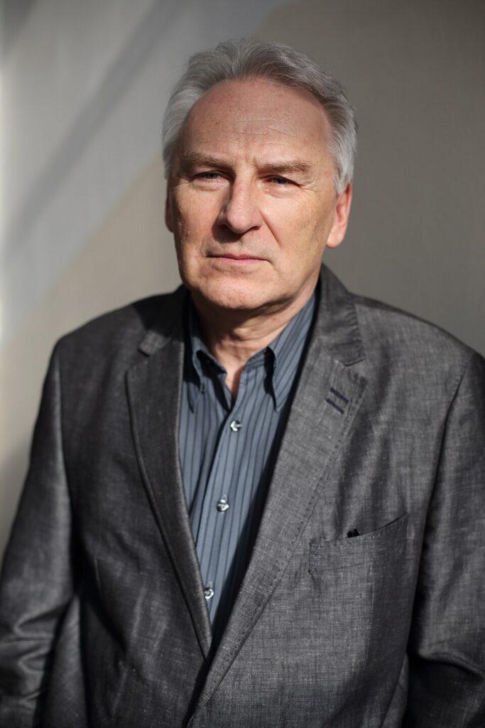 Mężczyzna w szarym garniturze stoi na tle jasnej ściany, z poważnym wyrazem twarzy.