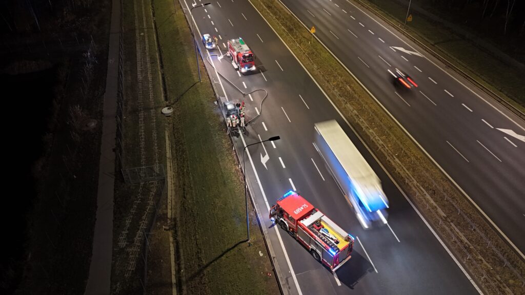 Strażacy gaszą pożar samochodu na poboczu drogi, obok stacji wozu strażackiego z włączonymi sygnałami.