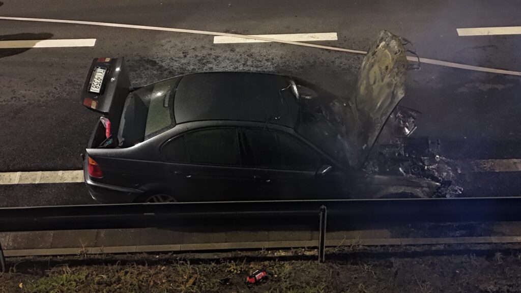 Czarny samochód osobowy z otwartą maską stoi na poboczu drogi, obok widoczne ślady uszkodzeń.