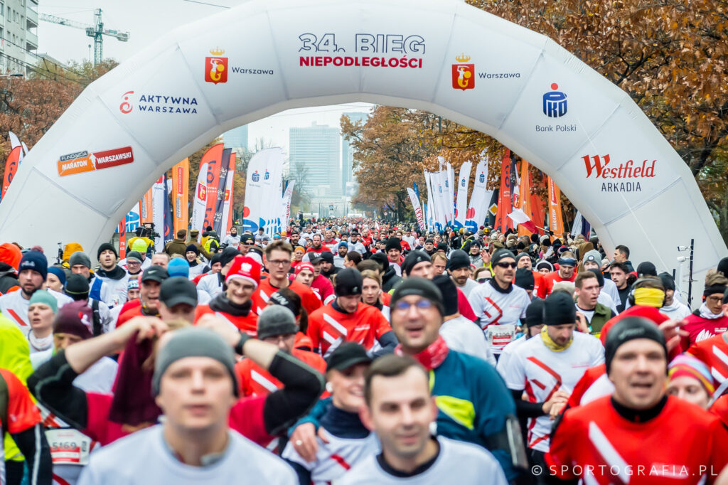 Uczestnicy biegu w czerwonych i białych koszulkach startują pod dużym łukiem z napisem "34. Bieg Niepodległości".