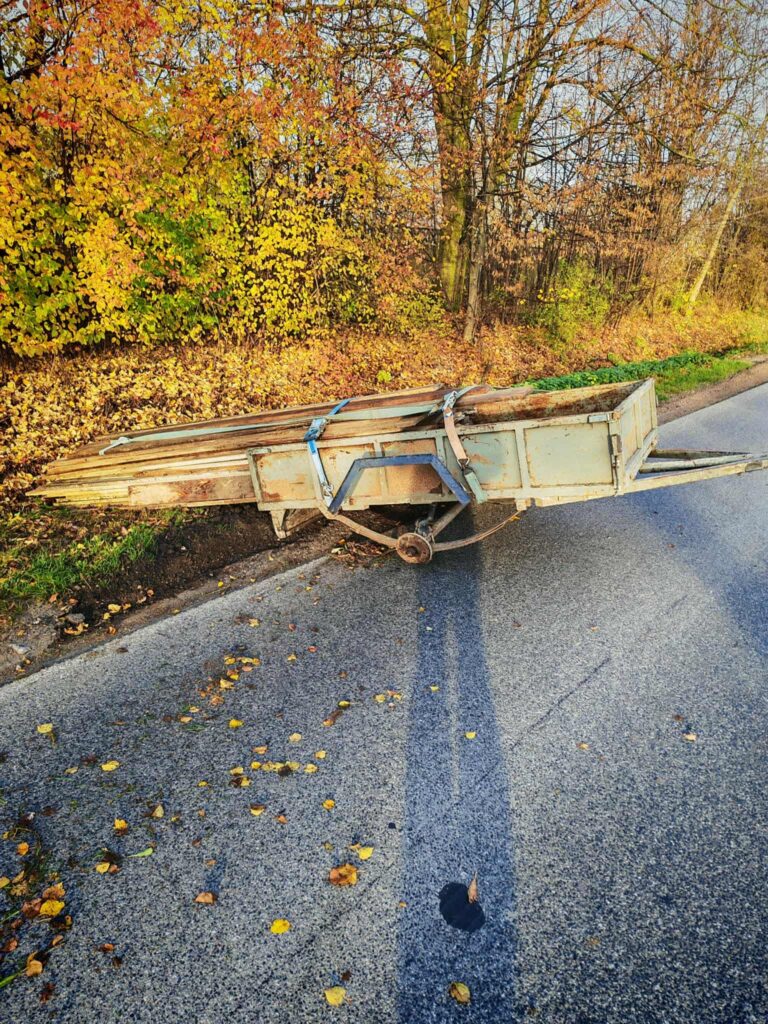 Zardzewiała przyczepa z drewnem stoi na poboczu drogi, otoczona kolorowymi liśćmi jesieni.