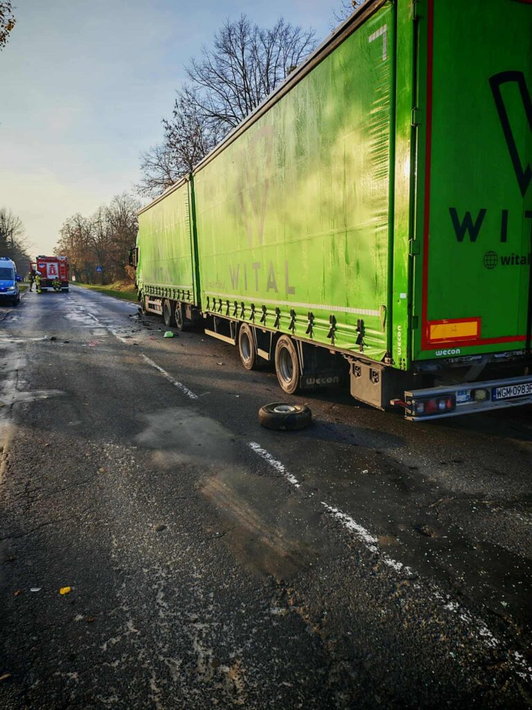 Zielona ciężarówka z uszkodzoną oponą zatrzymana na drodze, obok widać pojazd ratunkowy oraz służby.