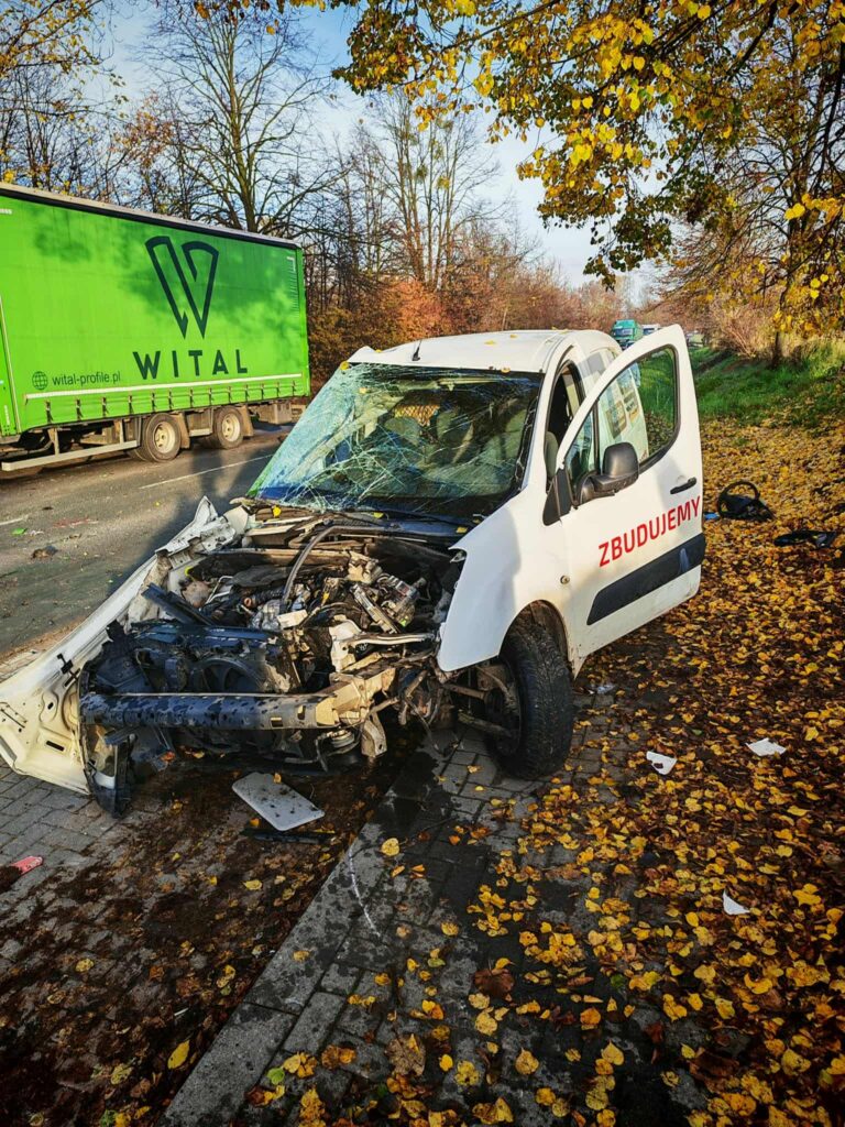 Rozbity samochód dostawczy z uszkodzoną przednią częścią stoi na drodze, obok przejeżdża zielona ciężarówka.