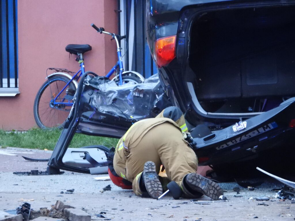Strażak w mundurze bada uszkodzone wnętrze przewróconego samochodu na ulicy, obok stoi niebieski rower.