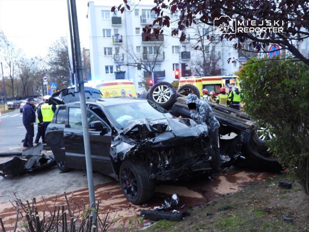 Wielokrotny wypadek drogowy z uszkodzonym czarnym SUV-em, obok interweniujący policjanci i ratownicy medyczni.
