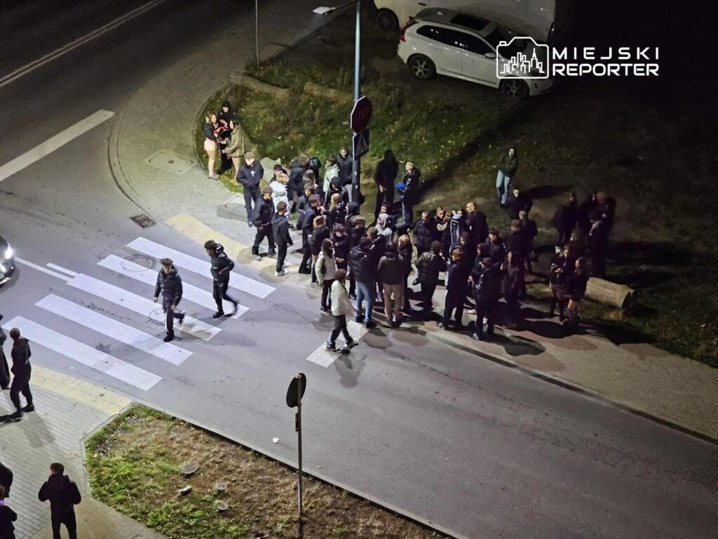 Grupa ludzi w ciemnych ubraniach gromadzi się na przejściu dla pieszych w nocy, obok zaparkowanych samochodów.