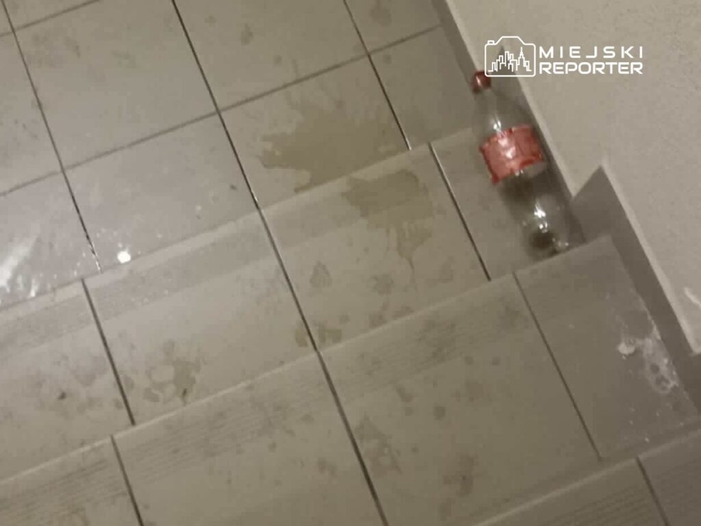 Na schodach widać rozlaną wodę oraz pustą butelkę po napoju. Podłoże pokryte plamami.