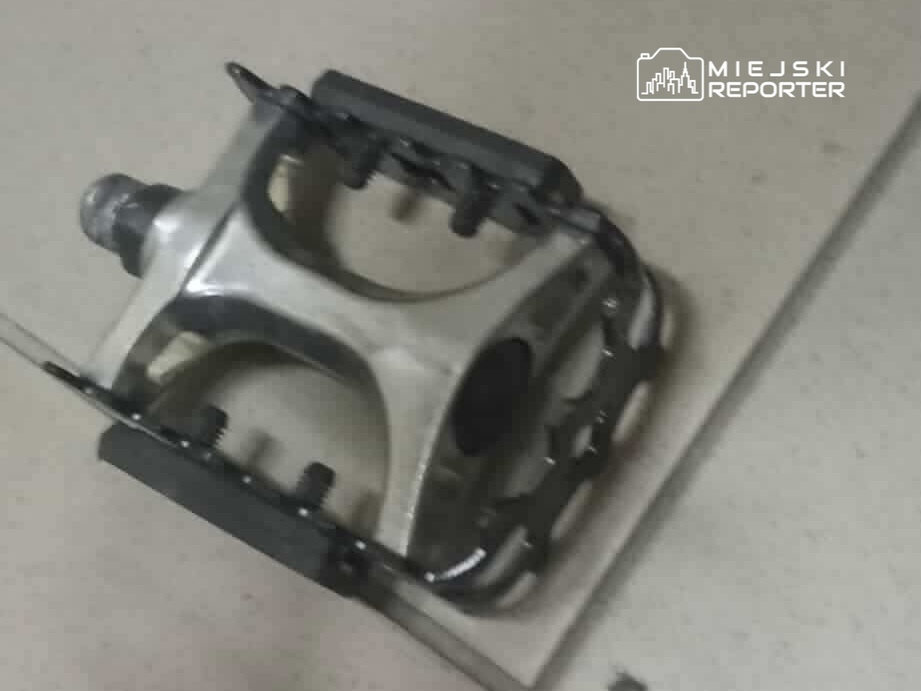 Metalowy pedał rowerowy leży na jasnej powierzchni, z widocznymi elementami mocującymi.