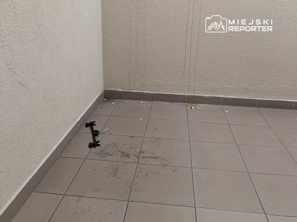 Pusty korytarz z brudną posadzką, na której leży czarny statyw i resztki śmieci. Ściany gładkie, jasne.