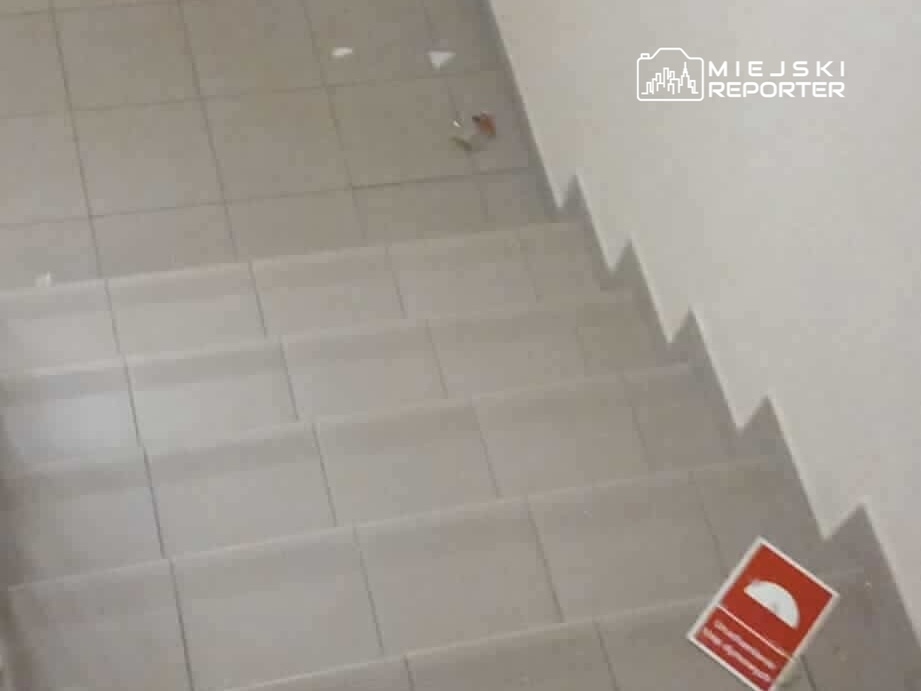 Na schodach widoczny jest czerwony znak ostrzegawczy oraz kilka drobnych odpadków na podłodze.