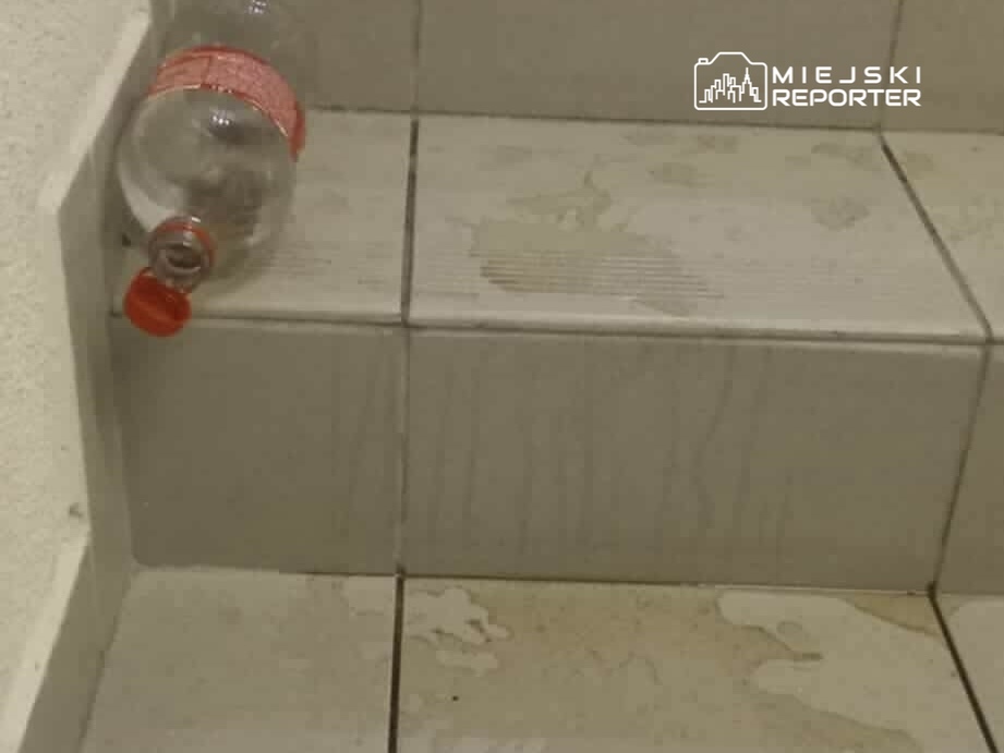 Na schodach leży plastikowa butelka z czerwoną nakrętką, obok widoczne plamy wody na kafelkach.