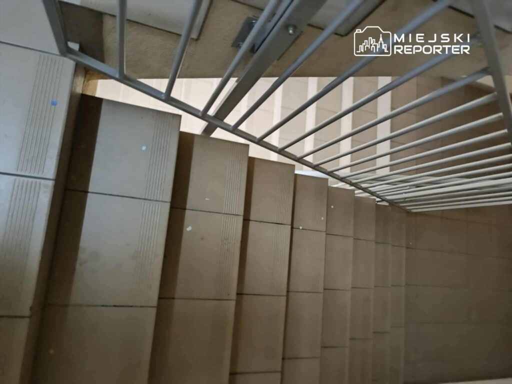 Schody z jasnymi płytkami prowadzące w dół, z widoczną metalową barierką na górze.