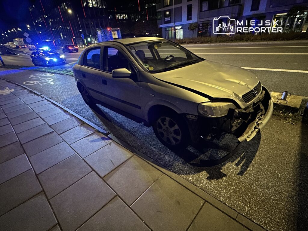 Uszkodzony samochód osobowy zaparkowany na chodniku, obok widać policyjny radiowóz z włączonymi sygnałami świetlnymi.