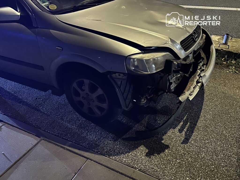 Uszkodzony samochód osobowy z zniszczonym przodem stoi na ulicy nocą, obok widoczna kostka brukowa.