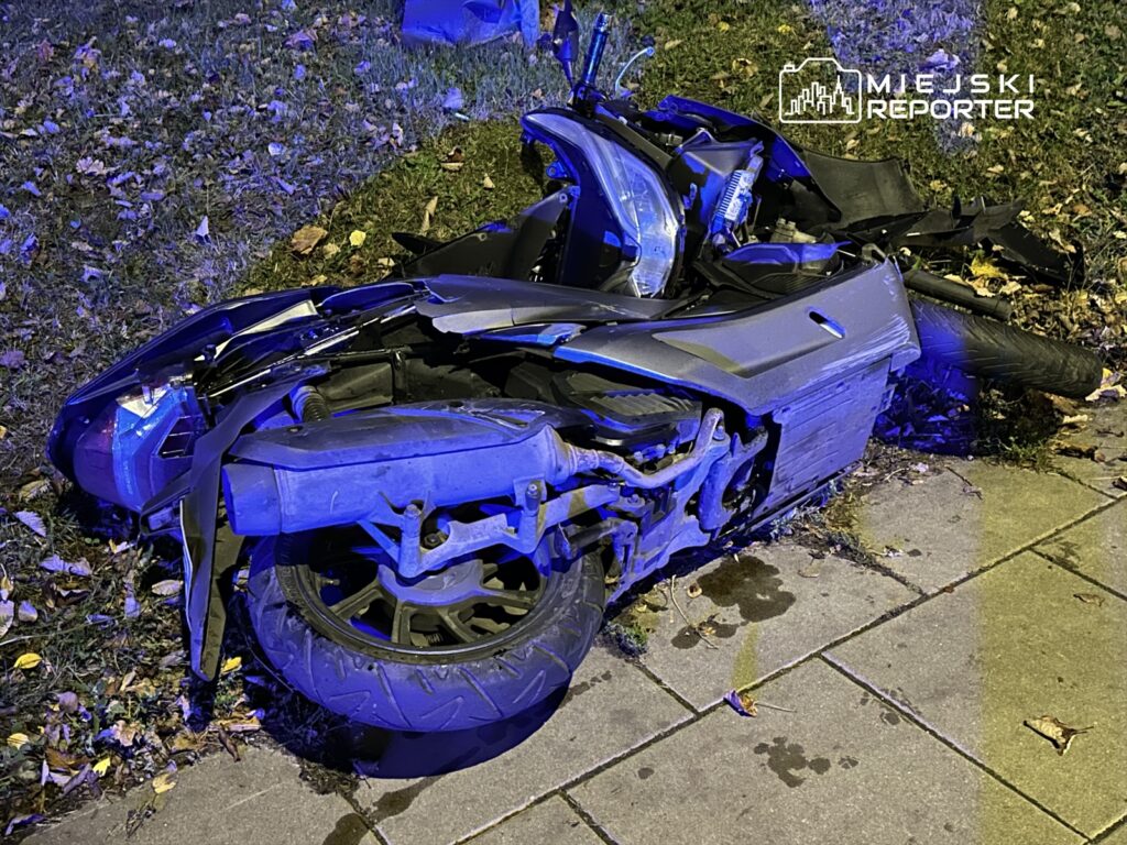 Uszkodzony motocykl leży na trawie obok chodnika, otoczony opadłymi liśćmi, w blasku niebieskich świateł.