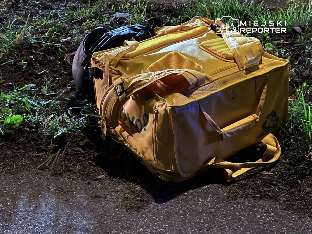 Żółta torba leży na poboczu drogi, otoczona trawą i liśćmi, w nocy oświetlona niebieskim światłem.