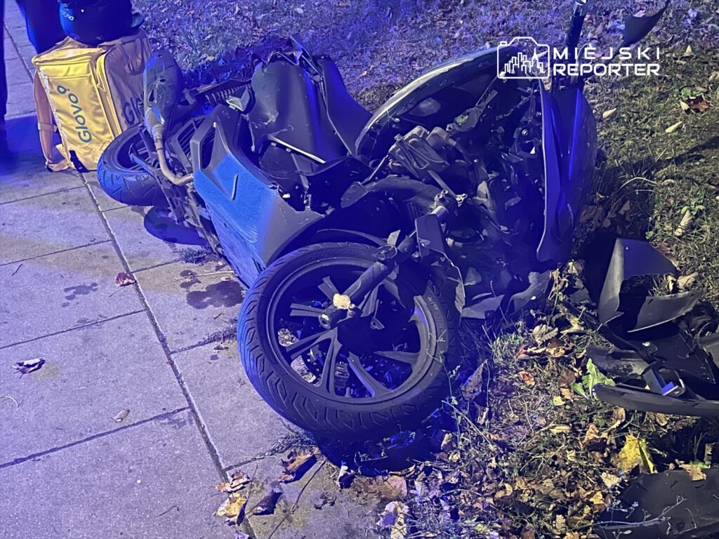 Zniszczony motocykl leży na chodniku, obok torba dostawcza. W tle widać zieleń i liście na ziemi.