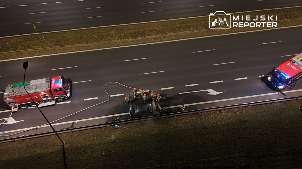 Grupa strażaków w kaskach pracuje nad usunięciem uszkodzonego pojazdu na autostradzie, obok dwóch wozów strażackich.