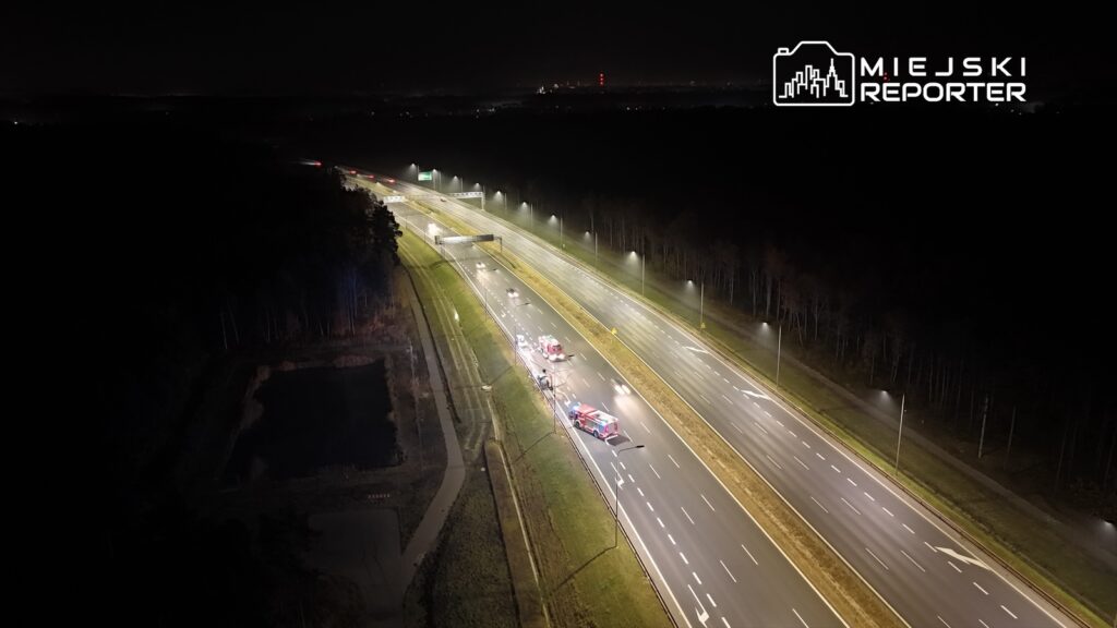 Na drodze nocą, widać kilka wozów strażackich zatrzymanych obok jezdni, otoczonych przez oświetlenie uliczne.